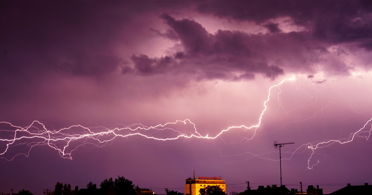 Un fulger ramificat se întinde pe orizontală pe un cer acoperit de nori în nuanțe de violet, deasupra unui oraș.