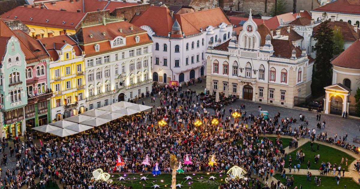 Imagine aeriană din Piața Unirii din Timișoara, unde sute de oameni sunt adunați în fața clădirilor istorice colorate. În prim-plan se vede zona verde a pieței, iar în jur apar fațade baroce, acoperișuri roșii și clădiri emblematice din centrul orașului.