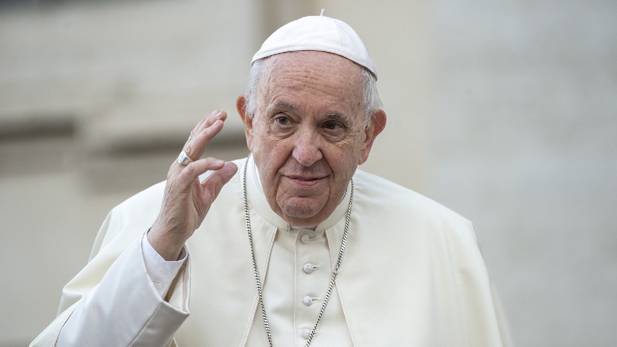În imagine este ilustrat Papa Francisc, în timp ce ține mâna dreaptă ridicată