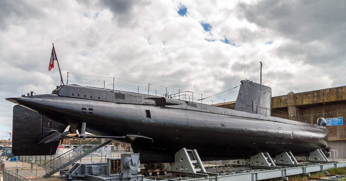 Imaginea prezintă un submarin francez expus pe uscat, așezat pe o structură metalică de suport. Nava, vopsită în negru, este masivă și păstrează detalii militare ale designului său din perioada postbelică. În fundal se observă o bază navală și drapelul Franței, iar cerul înnorat accentuează atmosfera istorică și solemnă a scenei.