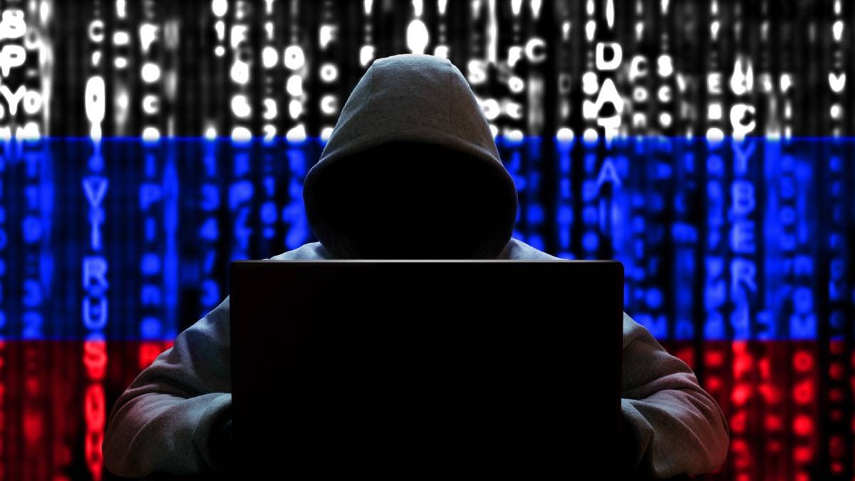 Un spion hacker îmbrăcat în hanorac și cu laptopul în brațe în timp ce pe fundal se vede steagul Rusiei.