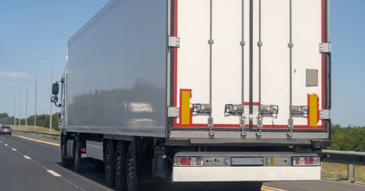 Imaginea arată partea din spate a unui camion de mare tonaj, aflat pe o autostradă. Drumul este asfaltat, cu mai multe benzi pe sens