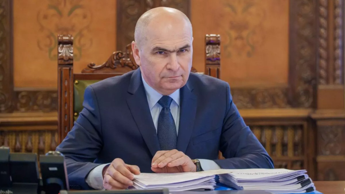 În imagine este ilustrat Ilie Bolojan, îmbrăcat în costum albastru închis. Acesta stă la birou.