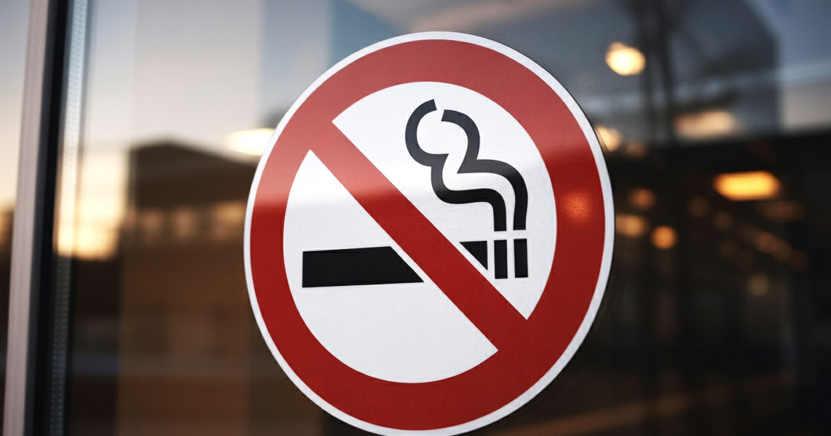 Imaginea prezintă de aproape un autocolant rotund de avertizare, de culoare roșu și alb, aplicat pe o suprafață de sticlă, care semnalează interzicerea fumatului. Semnul conține o țigară stilizată din care iese fum, barată de o linie diagonală roșie, indicând interdicția.