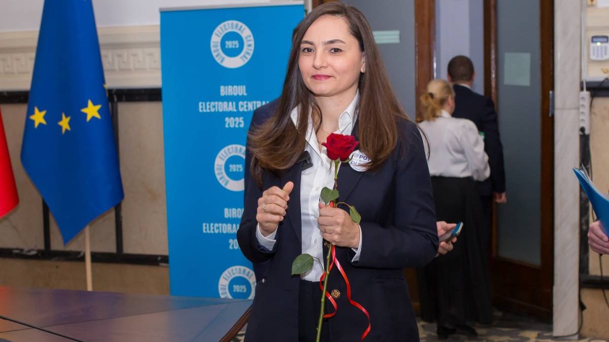 Anamaria Gavrilă îmbrăcată într-un costum negru și cămașă albă, în timpul ce ține un trandafir roșu în mână, surprinsă veselă la Biroul Electoral Central.