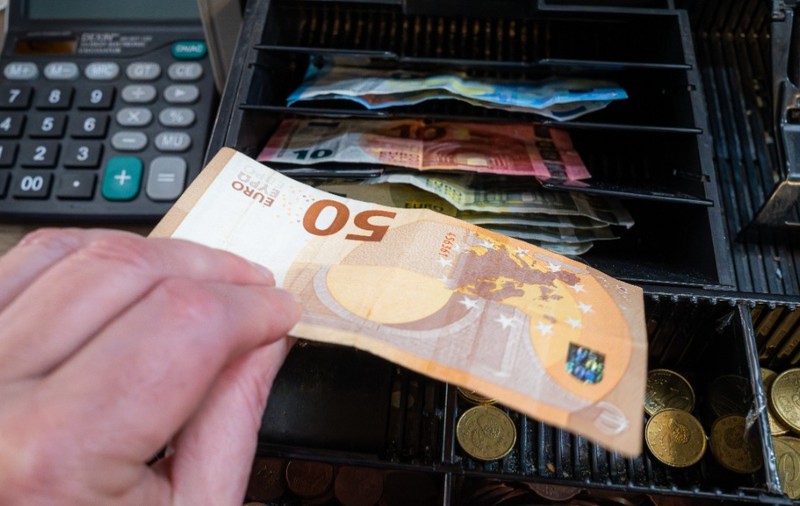 O mână care pune o bancnotă de 50 de euro într-un sertar deschis al unei case de marcat, lângă compartimentele pline cu monede și bancnote euro.