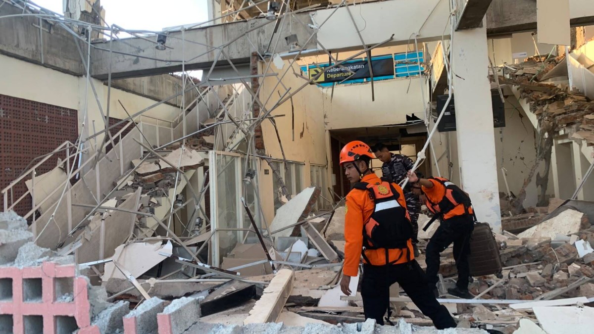 Imaginea înfățișează salvatori în uniforme portocalii de protecție care caută printre dărâmăturile și molozul unei clădiri prăbușite în urma unui cutremur.