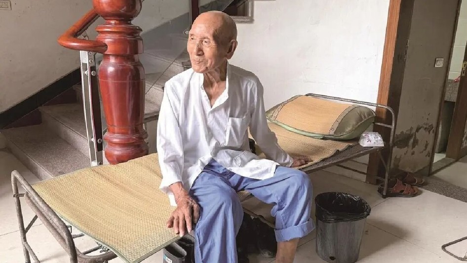 Un bărbat de 100 de ani din China stă pe un pat pliant, poarta o bluza alba si pantaloni albastri