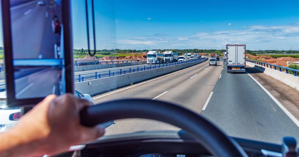 Un șofer de TIR la volanul camionului, conduce pe o autostradă într-o zi însorită, se vede că pe drum mai sunt și alte mașini.