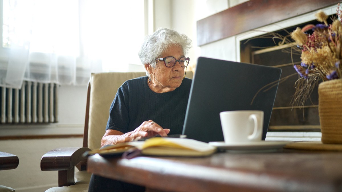 O pensionară cu părul cărut, în timp ce stă la un birou din sufragerie, în fața unui laptop și plătește facturi
