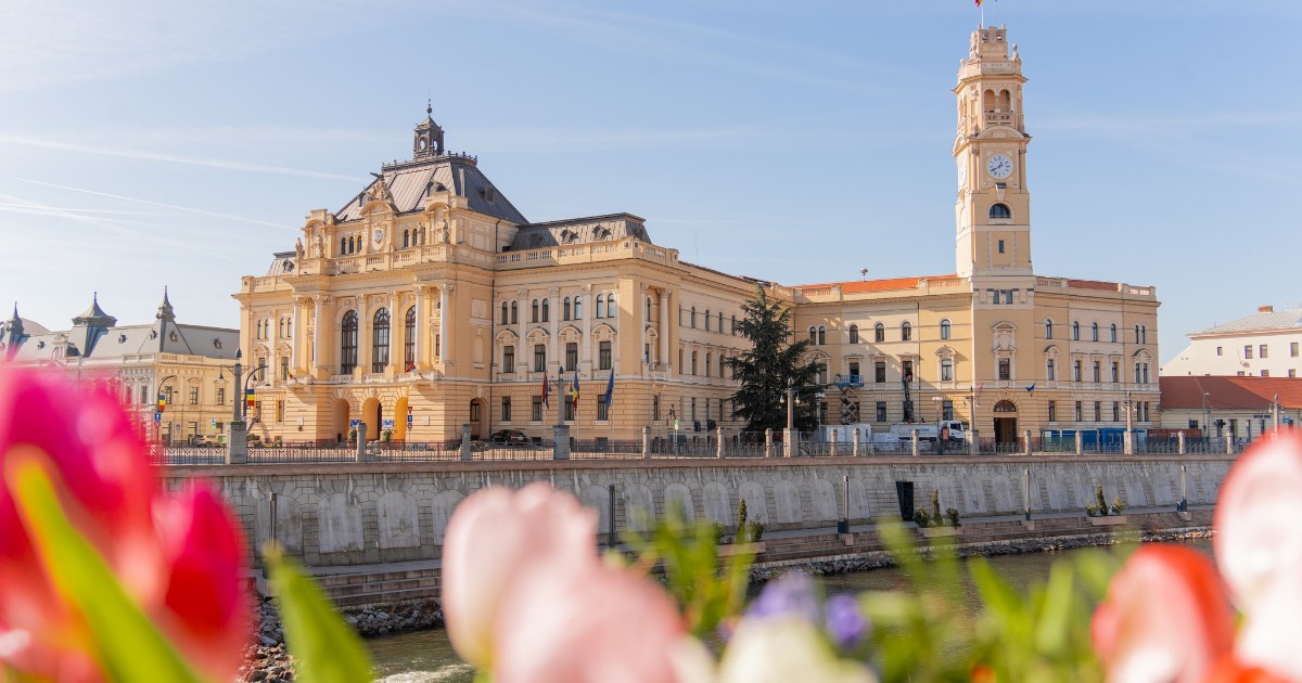 Clădirea impunătoare a Primăriei Oradea se reflectă spectaculos pe malul Crișului Repede, într-un peisaj urban elegant și luminos. În prim-plan, lalelele colorate oferă imaginii un aer de primăvară și pun în valoare arhitectura istorică a clădirii.