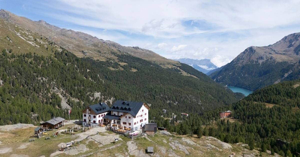 Imaginea surprinde o panoramă montană, cu o cabană albă situată pe o culme stâncoasă, înconjurată de pante împădurite și oferind o priveliște spre o vale adâncă unde se zărește un lac turcoaz.