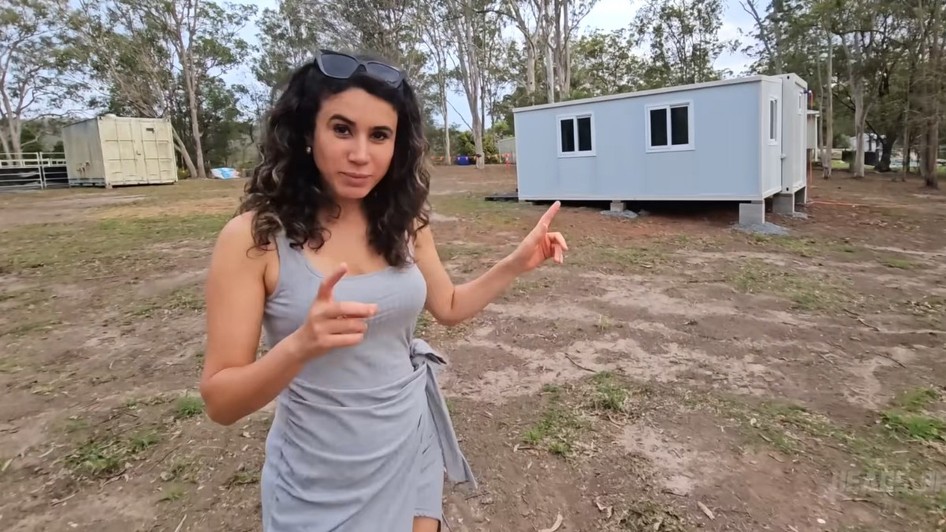 O femeie îmbrăcată într-o rochie gri care arată spre o casă modulară prefabricată, de culoare deschisă, amplasată într-un spațiu exterior deschis.