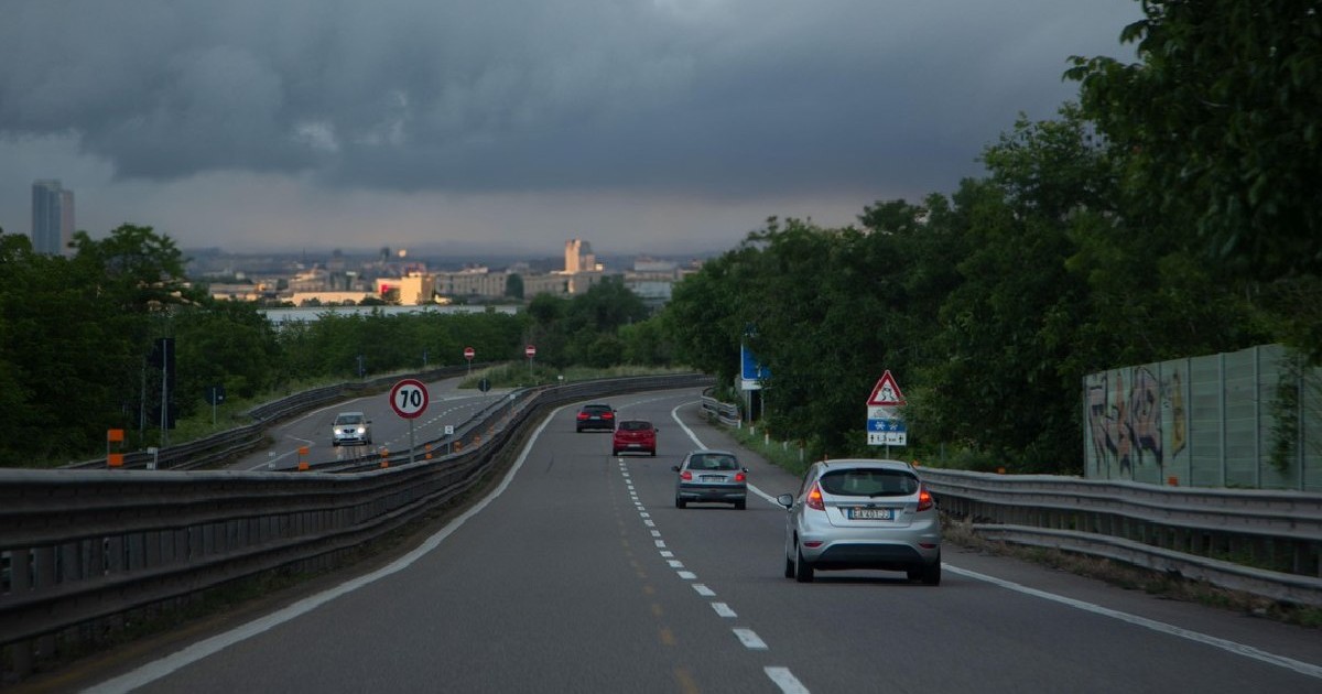Mai multe mașini circulă pe un drum cu două benzi, cerul este înnorat
