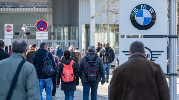 Un grup de persoane mergând spre intrarea unei facilități industriale, în dreapta fiind vizibile panouri cu siglele mărcii BMW și MINI.