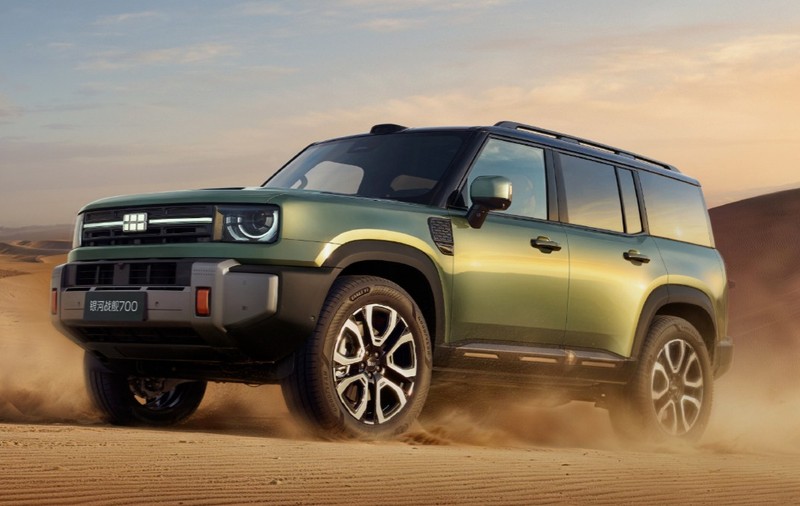 Un SUV off-road, de culoare verde măsliniu, rulând prin dunele de nisip ale unui deșert la apus.