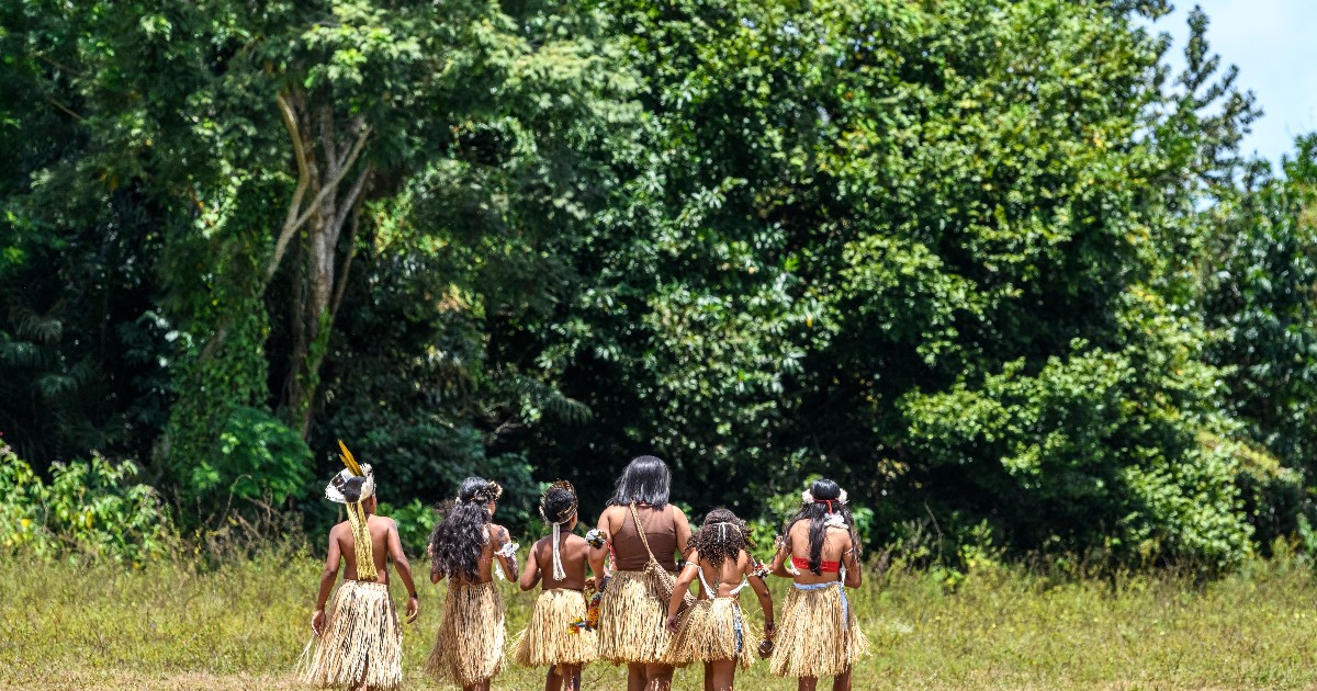 În imagine apare un grup de persoane îmbrăcate în costume tradiționale, care traversează o zonă de iarbă la marginea unei păduri tropicale. Costumele sunt decorate cu fuste din fibre vegetale, accesorii și elemente colorate, evidențiind specificul cultural al comunității.
