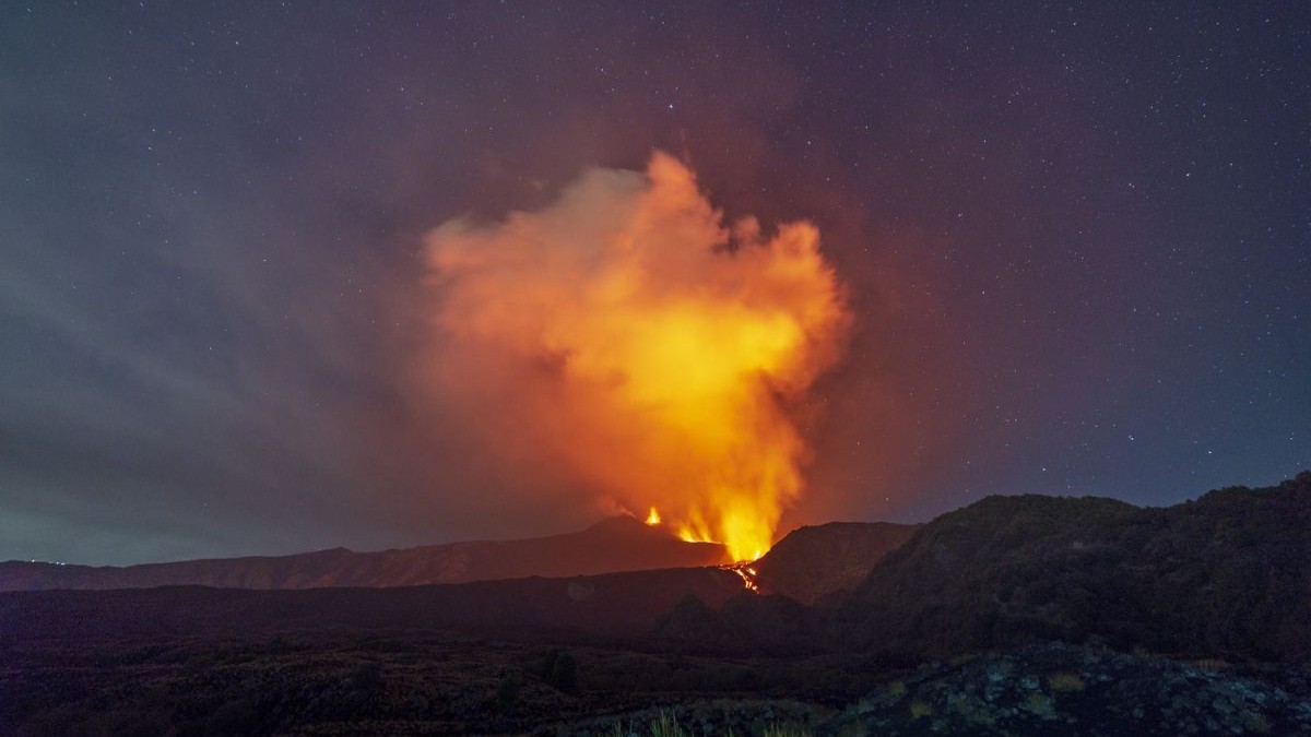 erupția vulcanului Etna din Italia, Sicilia, privit de la distanță. Din craterul situat pe creasta muntelui se înalță o coloană masivă și densă de fum, puternic luminată din interior de activitatea vulcanică. Pe versant se prelinge un râu de lavă incandescentă, iar în prim-plan se întinde un relief muntos accidentat, format din roci și vegetație măruntă.