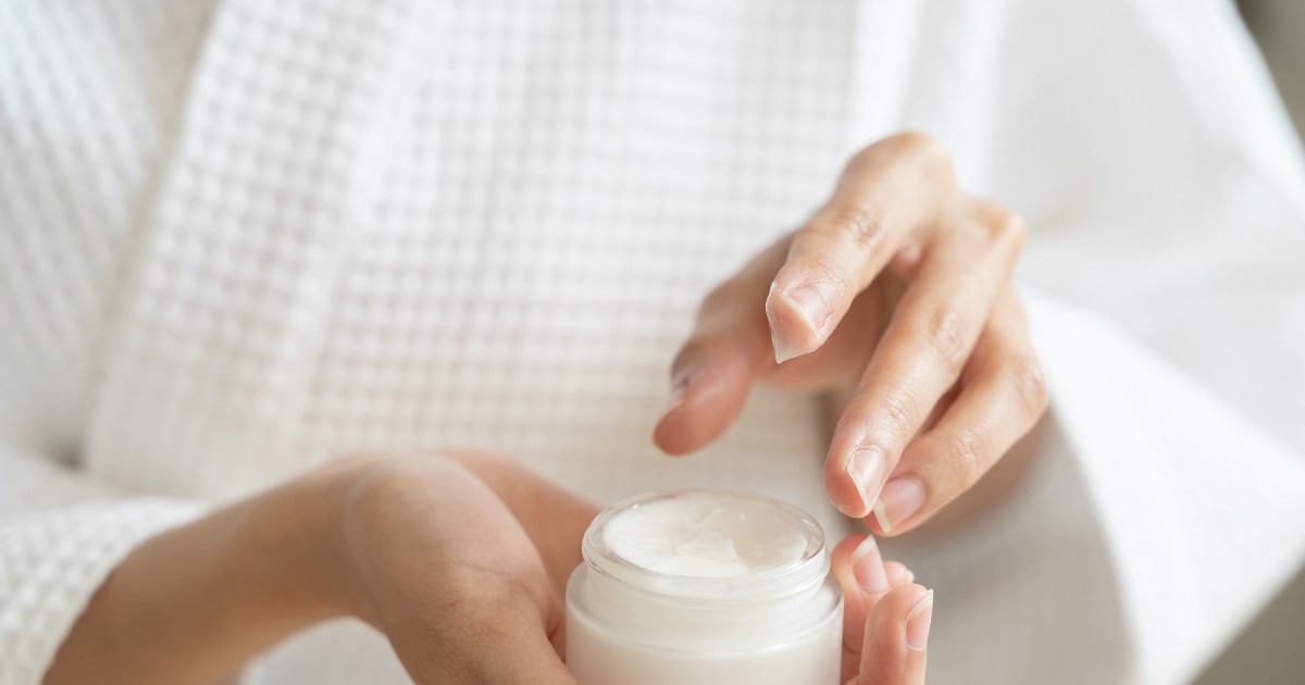 O persoană ține în palmă un recipient cu cremă cosmetică și se pregătește să aplice produsul pe piele. Imaginea sugerează o rutină de îngrijire personală și evidențiază utilizarea unui produs destinat hidratării și protecției pielii.