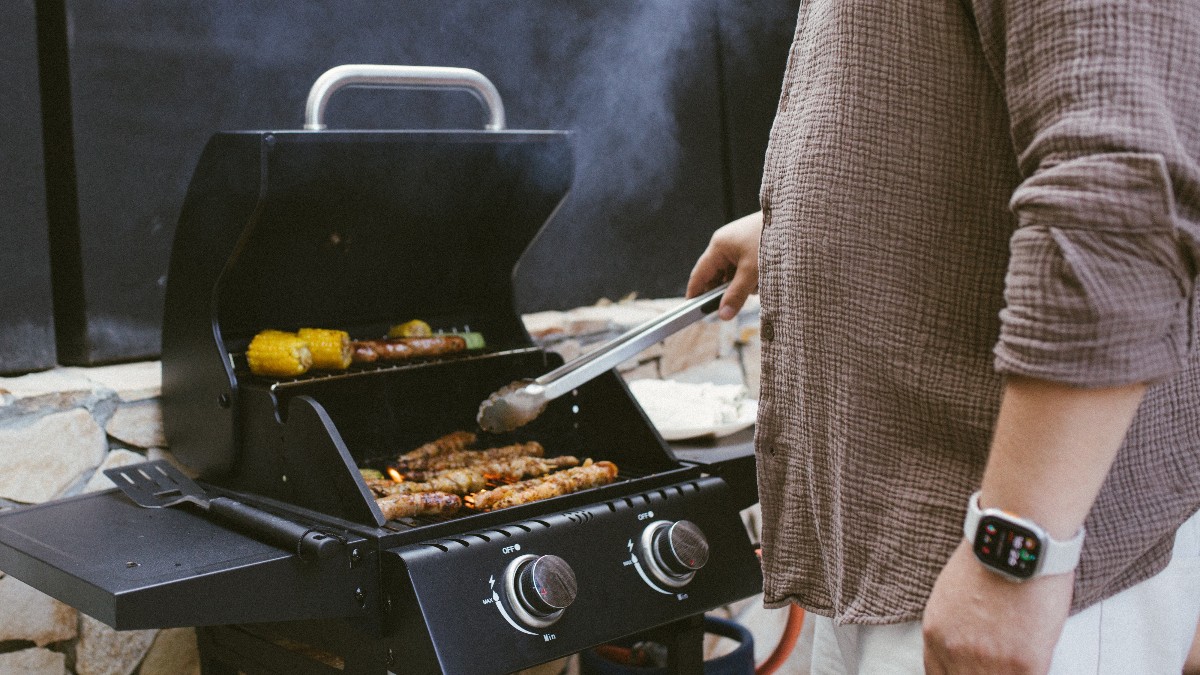 O persoană pregătește carne pe un grătar în aer liber, în timp ce fumul se ridică deasupra. Imaginea surprinde atmosfera relaxată a unei zile de primăvară, specifică ieșirilor de 1 Mai.