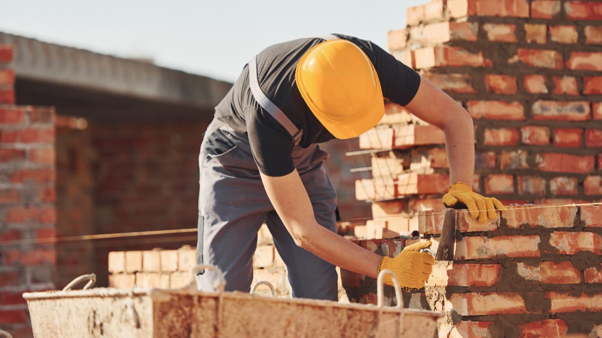 Un muncitor în construcții care poartă cască galbenă de protecție și mănuși, lucrând la zidirea unui perete din cărămidă roșie pe un șantier.