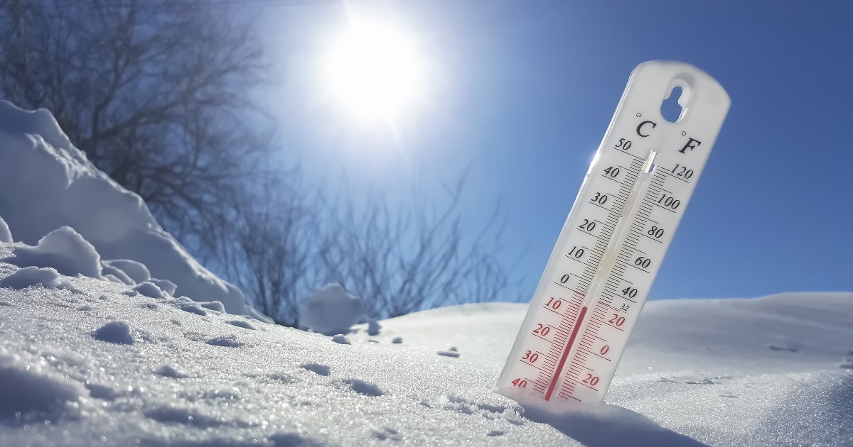 Imaginea prezintă un termometru alb înfipt în zăpadă sub un cer senin și însorit, indicând o temperatură scăzută.