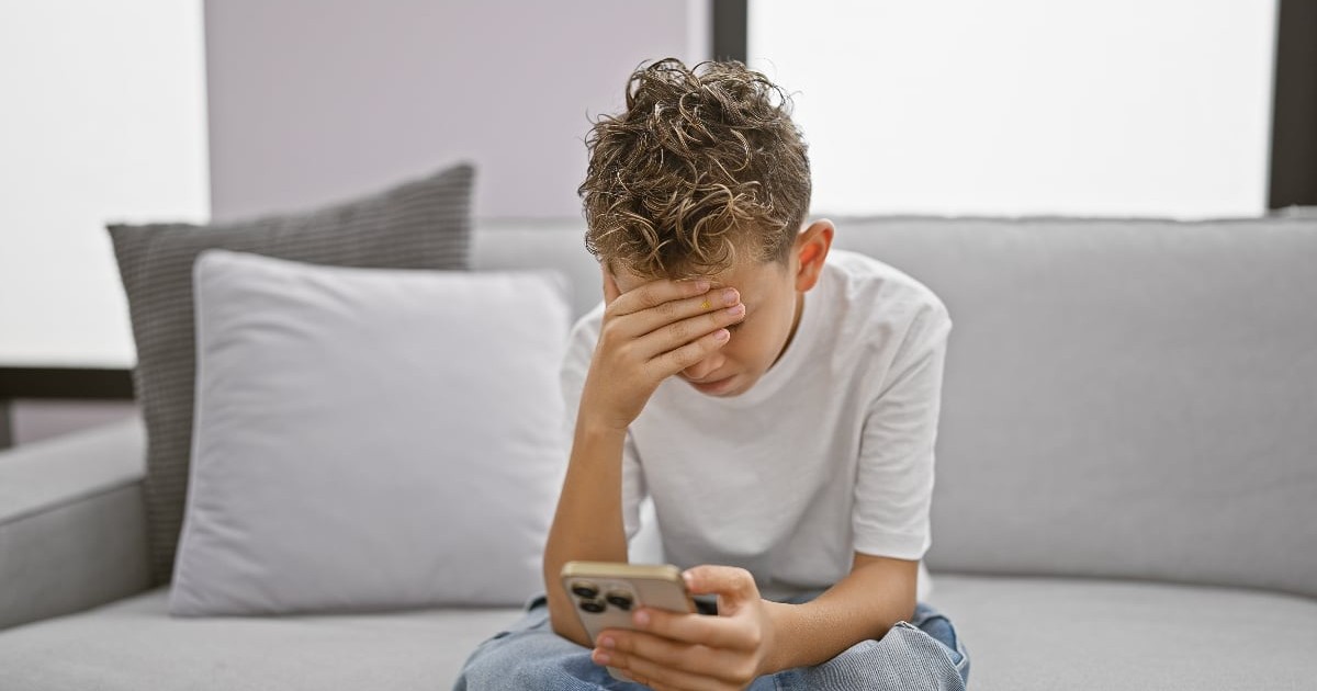 Imaginea prezintă un tânăr stând pe o canapea, ținând telefonul în mână și acoperindu-și fața cu cealaltă mână, semn de stres sau îngrijorare. Postura și expresia sugerează o stare de anxietate, posibil legată de conținutul pe care îl vede pe ecran.