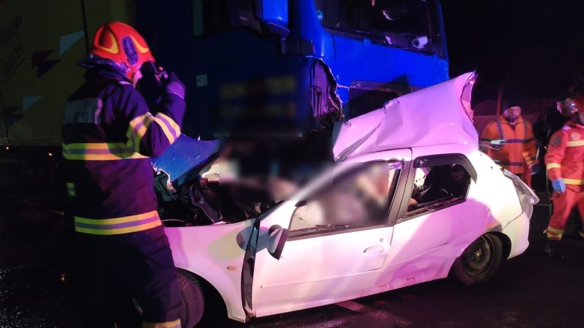Mașina de culoare albă, lovită în lateral de camionul de culoare albastră, în timp ce pompierii încearcă să scoată victimele din interior
