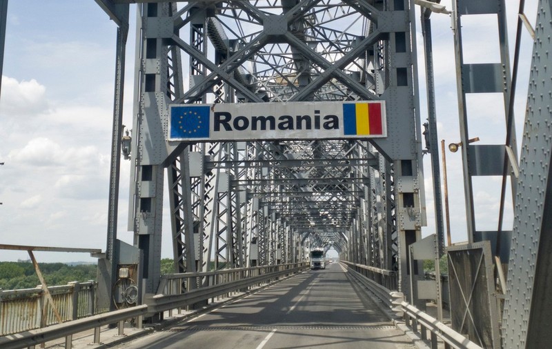 Podul Giurgiu-Ruse, podul Prieteniei, văzut dinspre Giurgiu. Pe șosea se află un TIR alb, iar deasupra ramei metalice ale podului se observă un semn de circulație cu textul „România”, steagul Uniunii Europene și steagul României