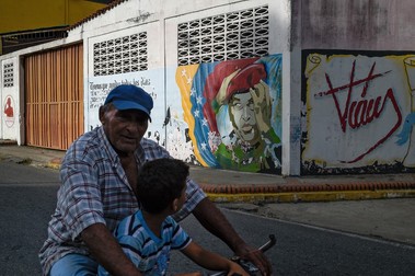 Un bărbat și un băiat merg cu bicicleta pe lângă o pictură murală care îl înfățișează pe fostul președinte venezuelean Hugo Chavez, în orașul său natal Sabaneta. Foto: Profimedia