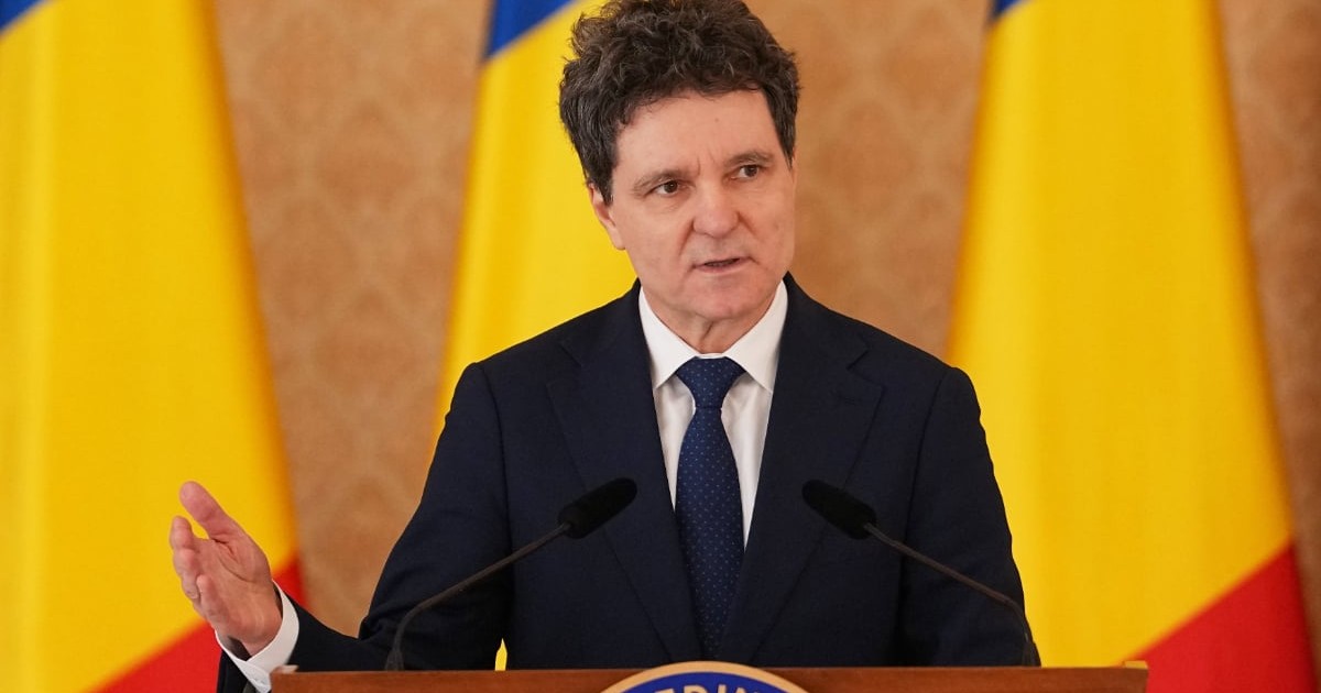Un bărbat îmbrăcat în costum închis la culoare vorbește la un pupitru oficial, având în față două microfoane. În fundal se văd steagurile României, iar postura lui sugerează că susține un discurs sau o declarație publică.