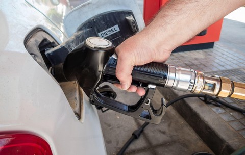 O mână care ține pistolul unei pompe de carburant introdus în rezervorul unui autoturism alb etichetat „DIESEL” într-o stație de alimentare.
