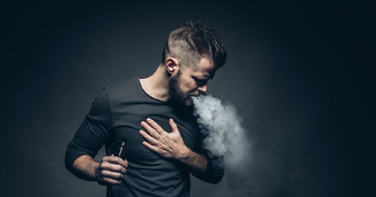 Un tânăr îmbrăcat în bluză neagră, pe fundal negru, vapează dintr-o tigară electronică- Vaporizarea e-lichidului aromatizat de la o țigară electronică