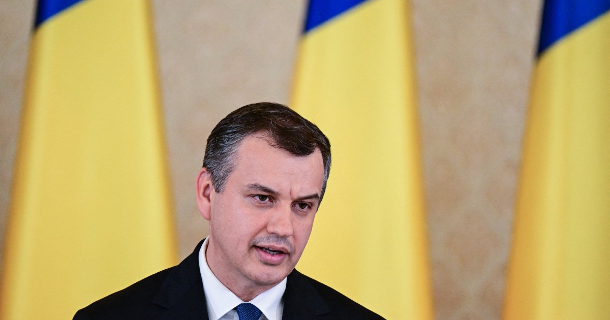 Imaginea îl surprinde pe Eugen Tomac în timpul unei declarații publice, îmbrăcat într-un costum închis la culoare și o cravată albastră. În fundal se disting drapele ale României, iar politicianul vorbește la un pupitru prevăzut cu microfoane.