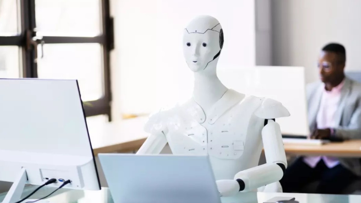 Un robot alb așezat la un birou, lucrând la un laptop.