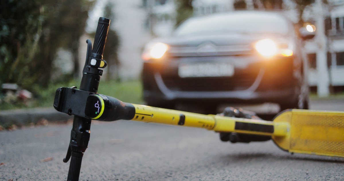 O trotinetă căzută pe asflat, după un accident rutier. În spatele este mașina care a lovit-o și are farurile aprinse
