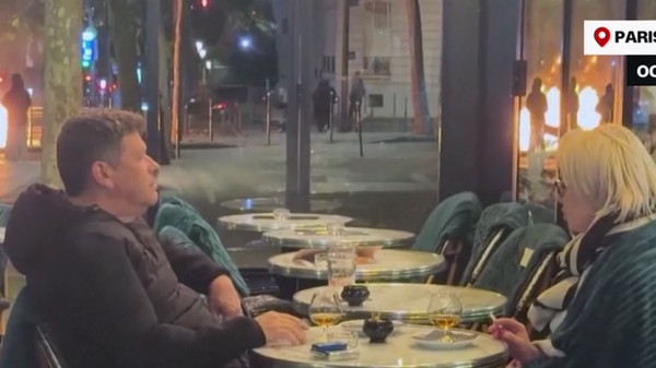 O imagine cu un cuplu care savurează o băutură la o terasă din Paris, lângă Arcul de Triumf, în timpul protestelor violente, a devenit virală. Sursă foto: Captură video/ Facebook /CBS News.