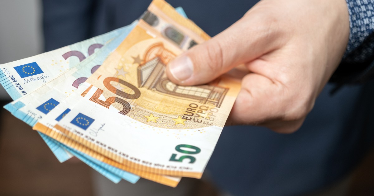 Imaginea este un prim-plan al unei mâini care ține un mănunchi de bancnote euro, cu accent pe cele de 50 de euro, care sunt cel mai vizibile. Persoana care ține bancnotele este îmbrăcată într-o cămașă cu model și un sacou închis la culoare.