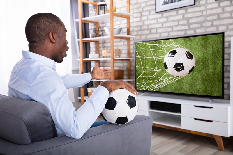 Shocked African Man Sitting On Sofa Watching Football Match On Television