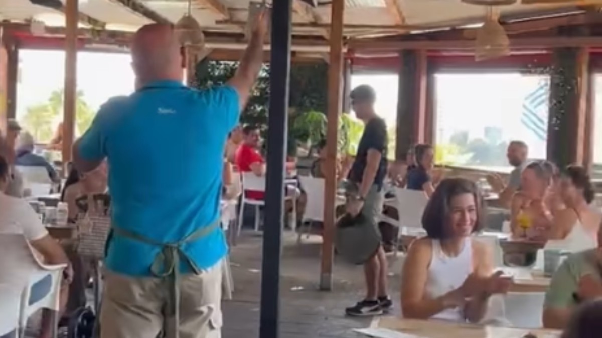 Un bărbat la un restaurant care poartă tricou albastru. Pe scaune se află oameni care aplaudă