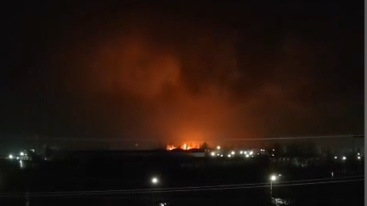 Imaginea surprinde o scenă nocturnă, dominată de un incendiu masiv vizibil la orizont, a cărui flacără roșiatică luminează intens cerul întunecat. În planul apropiat, se pot distinge siluetele unor clădiri și felinare stradale, care contrastează puternic cu lumina portocalie a focului din depărtare.