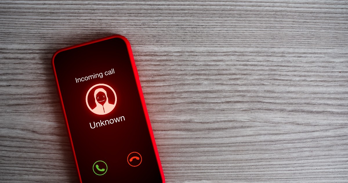 Imaginea arată un telefon mobil roșu, aflat pe o suprafață din lemn, afișând pe ecran un apel primit de la un număr necunoscut. Iconița ilustrată ca o figură cu glugă sugerează un potențial apel fraudulos, accentuând ideea de risc și înșelătorie.