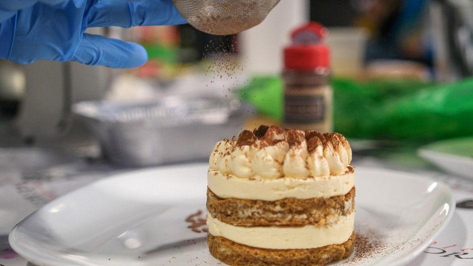 Tiramisu World Cup, un tiramisu construit sub forma de rotund, pe o farfurie. O persoană pune praf de cacao peste el