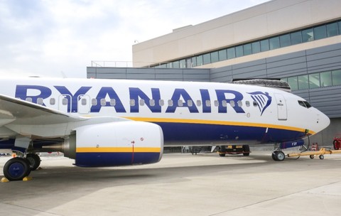 Un avion de pasageri al companiei Ryanair staționat pe pista unui aeroport, în fața unei clădiri moderne de terminal.