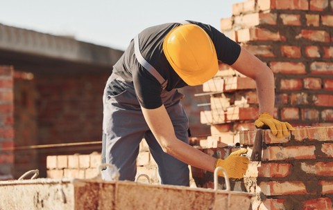 Un muncitor în construcții care poartă cască galbenă de protecție și mănuși, lucrând la zidirea unui perete din cărămidă roșie pe un șantier.
