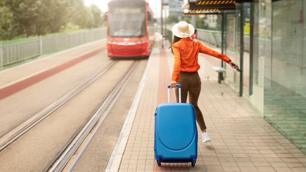 O femeie îmbrăcată într-o bluză portocalie și o pălărie de paie, trage după ea o valiză albastră mare, grăbindu-se pe peron. Ea aleargă în direcția unui tramvai roșu care se apropie sau pleacă pe șine, sugerând că se grăbește să îl prindă.
