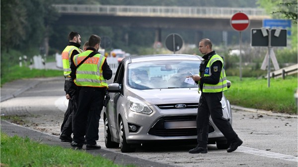 Polițiști germani purtând veste reflectorizante care efectuează un control de rutină asupra unui autoturism Ford argintiu oprit la marginea unui drum.
