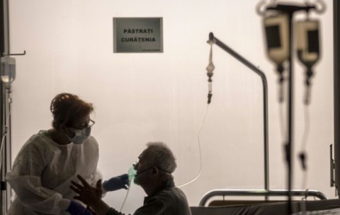 pacient stand pe pat intr-un salon de spital cu o perfuzie la capul patului. Langa el sta o femeie care-i pune masca de oxigen pe gura