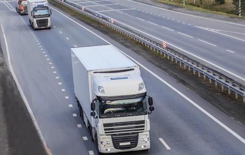 Un camion alb de mare tonaj circulând pe banda unei autostrăzi cu mai multe benzi, urmat la distanță de alte autovehicule comerciale.   