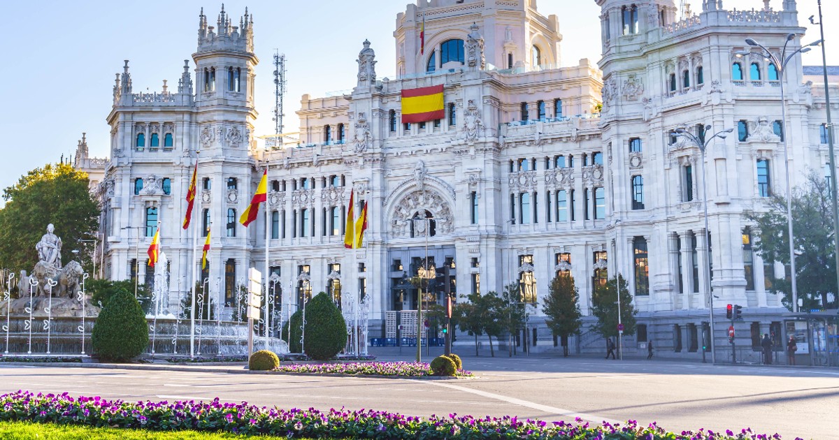 Imaginea prezintă impunătoarea clădire a Palatului Cibeles din Madrid, Spania, un edificiu cu arhitectură albă, neoclasică, deasupra căruia flutură steagul Spaniei. În prim-plan se observă o zonă verde cu flori violete și o fântână arteziană cu statui, încadrată de o stradă largă.